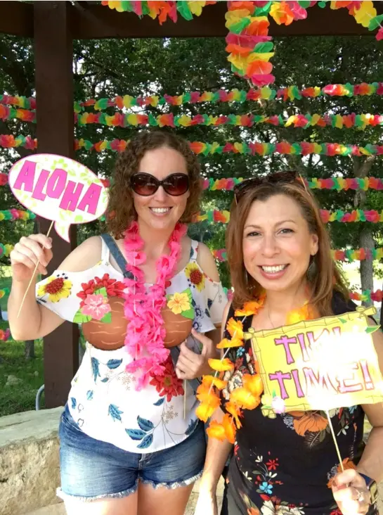 Real luau party — two women holding Aloha and Tiki Time photo props under flower garland arch