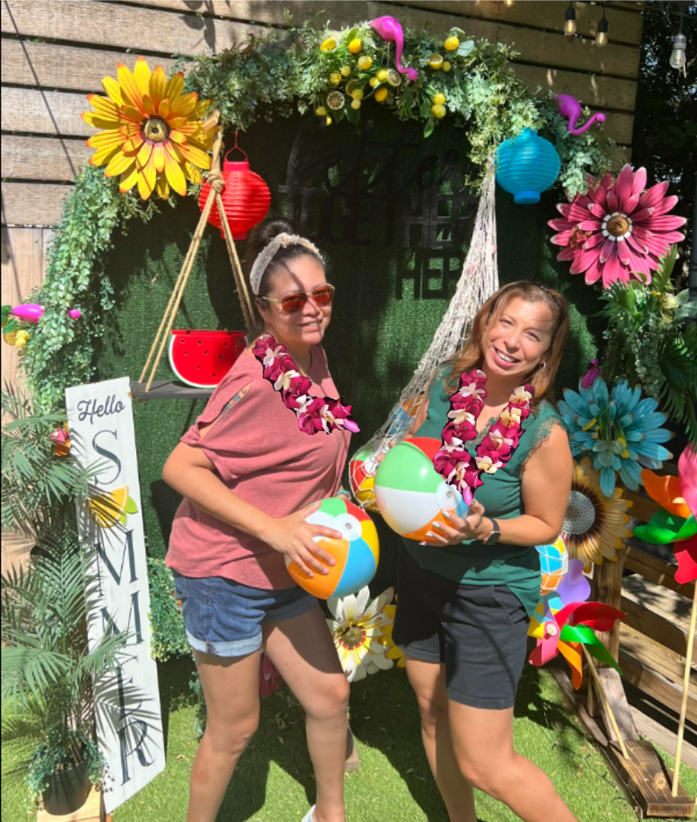 Real luau party — two women with leis and beach balls in front of a tropical floral photo arch