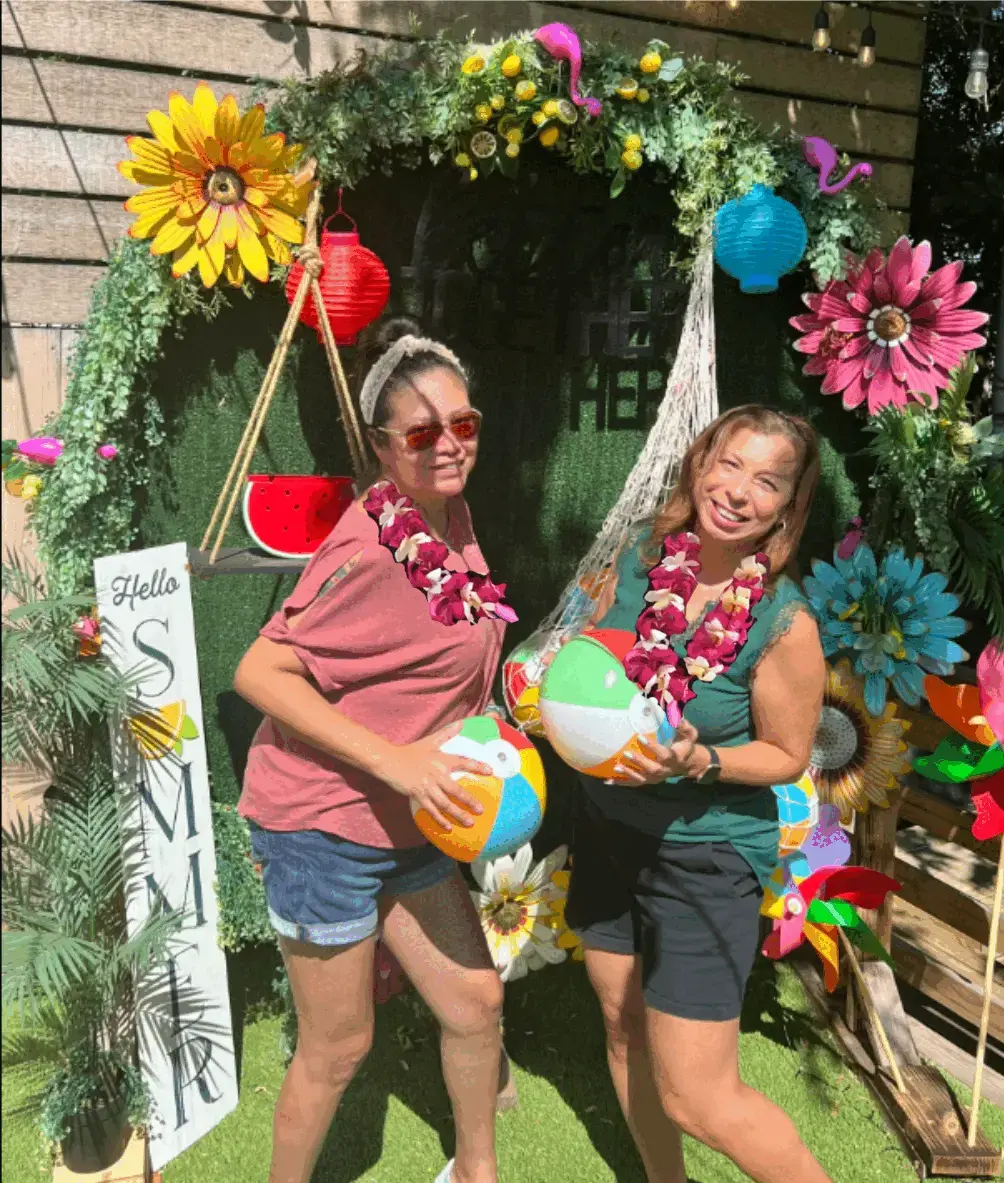 Real luau party — two women with leis and beach balls in front of a tropical floral photo arch