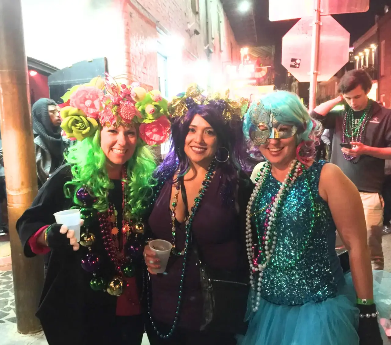 Real Mardi Gras party — three women in colorful wigs and beads on a New Orleans street at night