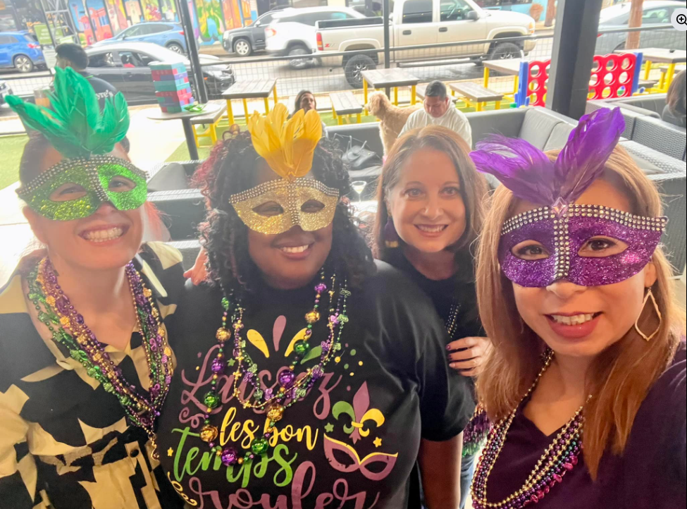 Real Mardi Gras party — four women wearing feathered masks and bead necklaces smiling at a daytime celebration