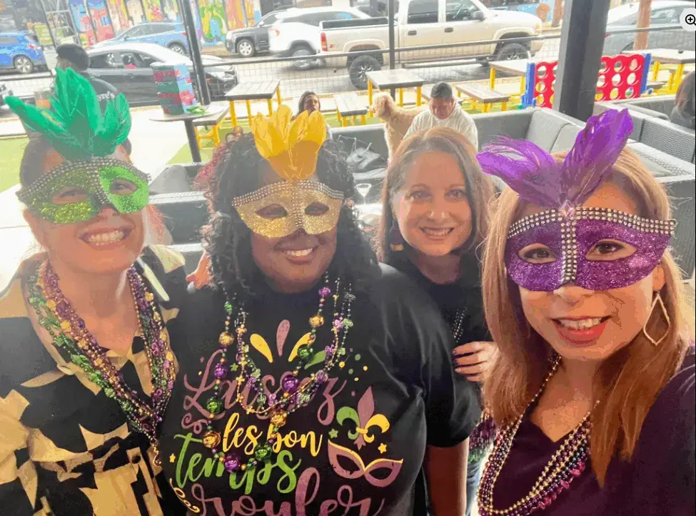 Real Mardi Gras party — four women wearing feathered masks and bead necklaces smiling at a daytime celebration
