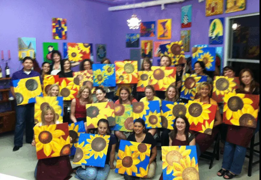 Real paint and sip party — large group holding sunflower paintings in a purple studio